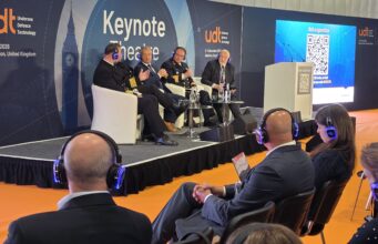 Panel discussion on stage with five men in suits and uniforms seated in white chairs at a UDT Keynote Theatre event, audience wearing blue headphones on orange floor.