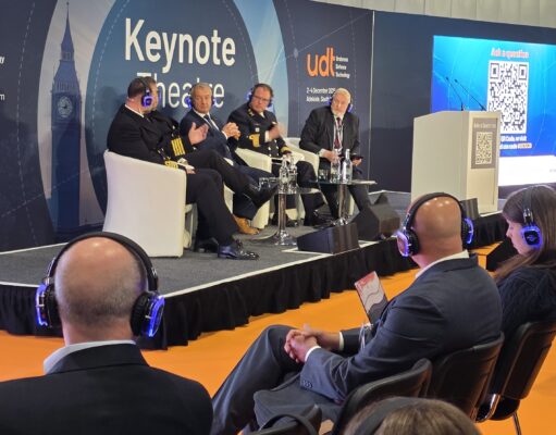 Panel discussion on stage with five men in suits and uniforms seated in white chairs at a UDT Keynote Theatre event, audience wearing blue headphones on orange floor.