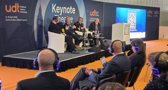 Panel discussion on stage with five men in suits and uniforms seated in white chairs at a UDT Keynote Theatre event, audience wearing blue headphones on orange floor.