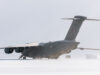 Large military transport aircraft (four engines) on a snow-covered tarmac with a ground crew member nearby.