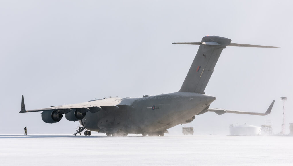 Large military transport aircraft (four engines) on a snow-covered tarmac with a ground crew member nearby.