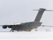 Large military transport aircraft (four engines) on a snow-covered tarmac with a ground crew member nearby.