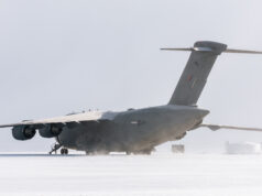 Large military transport aircraft (four engines) on a snow-covered tarmac with a ground crew member nearby.