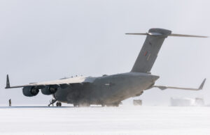 Large military transport aircraft (four engines) on a snow-covered tarmac with a ground crew member nearby.