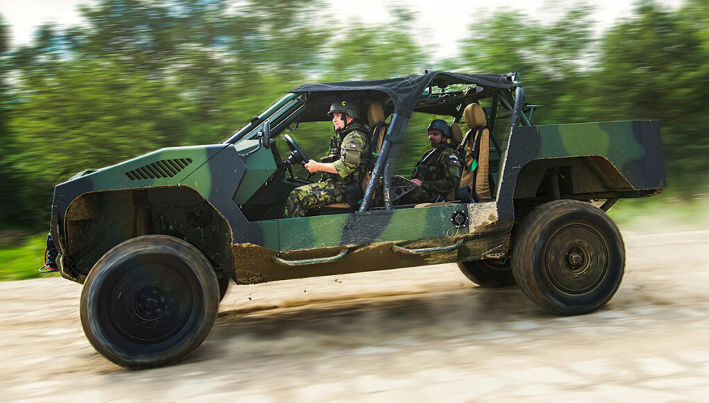 Two soldiers in camouflage drive a green open-top military vehicle along a dirt road, kicking up dust.