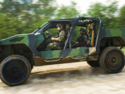 Two soldiers in camouflage drive a green open-top military vehicle along a dirt road, kicking up dust.