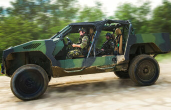 Two soldiers in camouflage drive a green open-top military vehicle along a dirt road, kicking up dust.