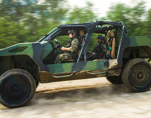 Two soldiers in camouflage drive a green open-top military vehicle along a dirt road, kicking up dust.
