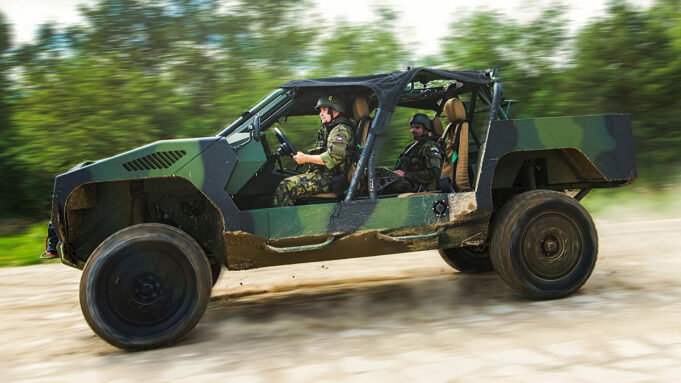 Two soldiers in camouflage drive a green open-top military vehicle along a dirt road, kicking up dust.