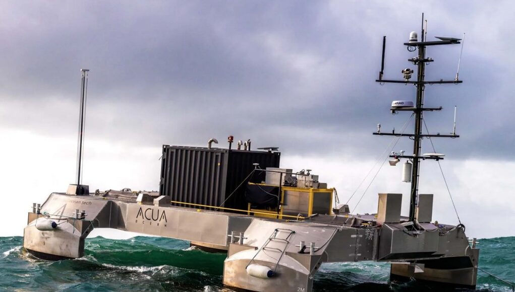 Metallic, rectangular research catamaran riding rough seas with a tall mast and lab equipment on deck.