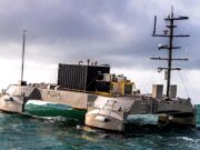 Metallic, rectangular research catamaran riding rough seas with a tall mast and lab equipment on deck.