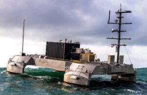 Metallic, rectangular research catamaran riding rough seas with a tall mast and lab equipment on deck.