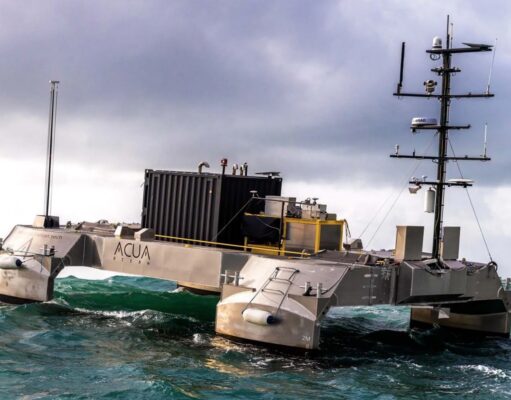 Metallic, rectangular research catamaran riding rough seas with a tall mast and lab equipment on deck.
