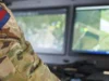 Soldier in camouflage uniform with a blue-and-red flag patch, working at a dual-monitor command station showing a map.