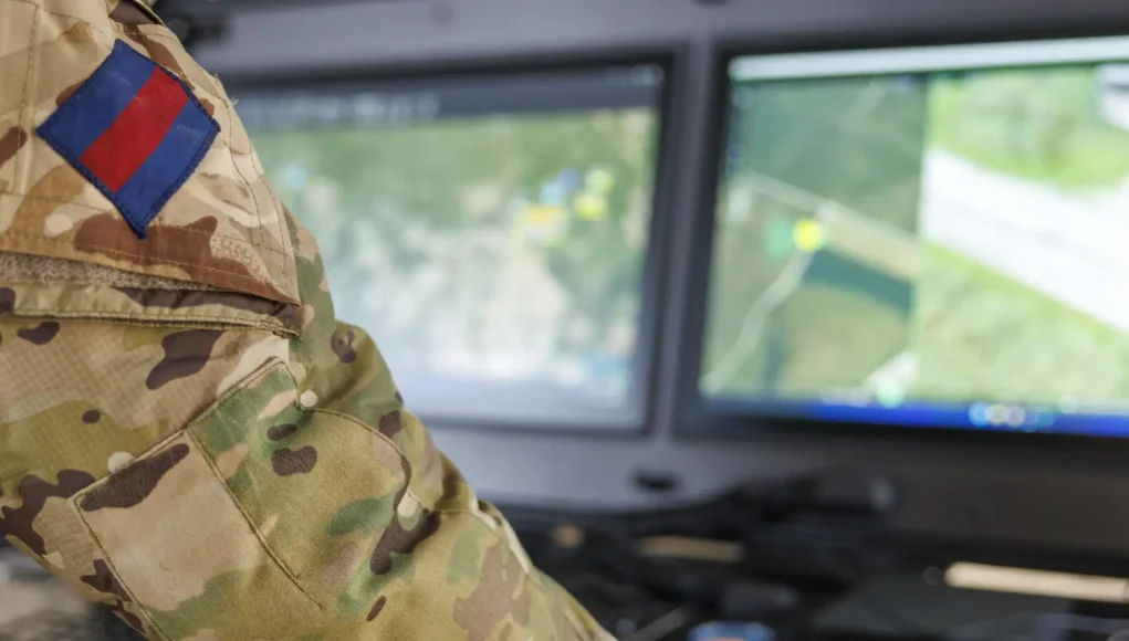 Soldier in camouflage uniform with a blue-and-red flag patch, working at a dual-monitor command station showing a map.