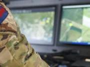 Soldier in camouflage uniform with a blue-and-red flag patch, working at a dual-monitor command station showing a map.