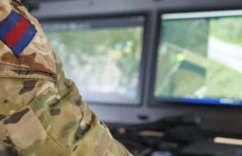 Soldier in camouflage uniform with a blue-and-red flag patch, working at a dual-monitor command station showing a map.