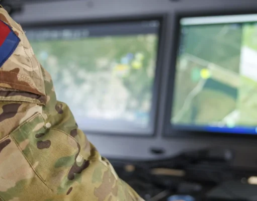 Soldier in camouflage uniform with a blue-and-red flag patch, working at a dual-monitor command station showing a map.