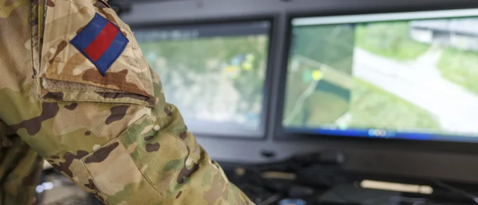 Soldier in camouflage uniform with a blue-and-red flag patch, working at a dual-monitor command station showing a map.