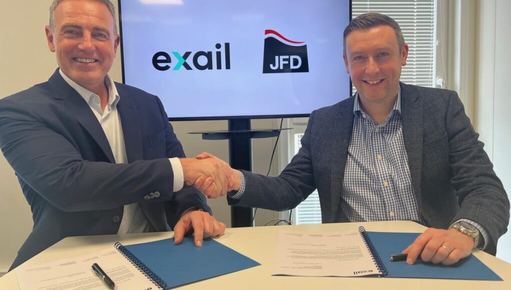 Two men in blazers shake hands over a table while signing documents, with Exail and JFD logos on screen behind them.