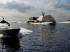 Large angular naval vessel gliding on calm sea with two smaller speedboats nearby under a cloudy sky, creating white wake.