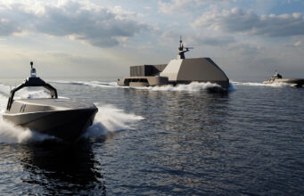 Large angular naval vessel gliding on calm sea with two smaller speedboats nearby under a cloudy sky, creating white wake.