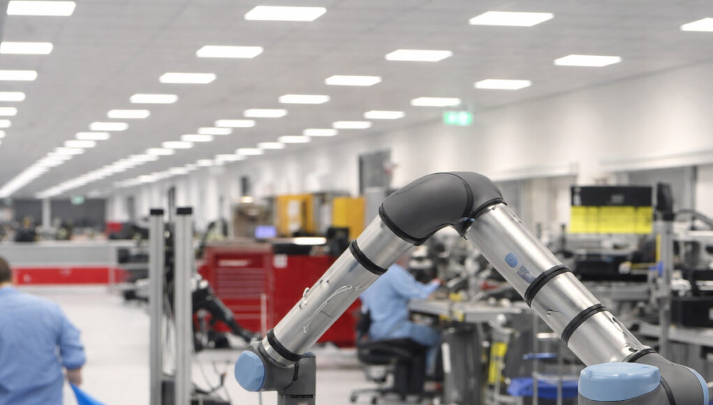 Industrial robotic arm on a production line with workers at benches in a bright factory.