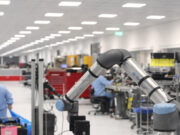 Industrial robotic arm on a production line with workers at benches in a bright factory.