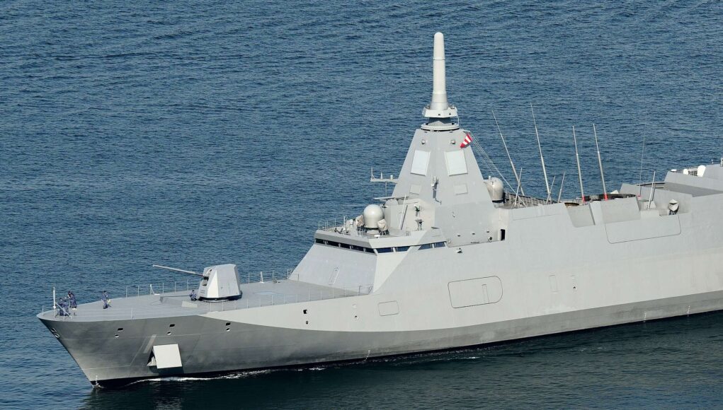 Large gray warship at sea with a tall central mast and sailors on the bow.