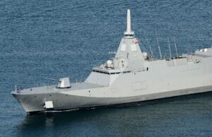 Large gray warship at sea with a tall central mast and sailors on the bow.