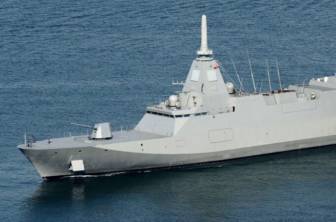 Large gray warship at sea with a tall central mast and sailors on the bow.