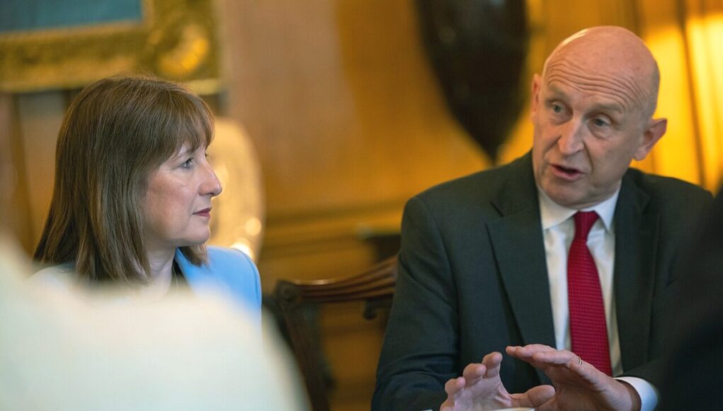 Two professionals sit at a table in a formal meeting, the man in a dark suit and red tie speaking while the woman in a light blue blazer listens.