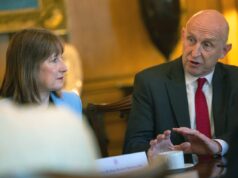 Two professionals sit at a table in a formal meeting, the man in a dark suit and red tie speaking while the woman in a light blue blazer listens.