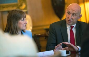 Two professionals sit at a table in a formal meeting, the man in a dark suit and red tie speaking while the woman in a light blue blazer listens.