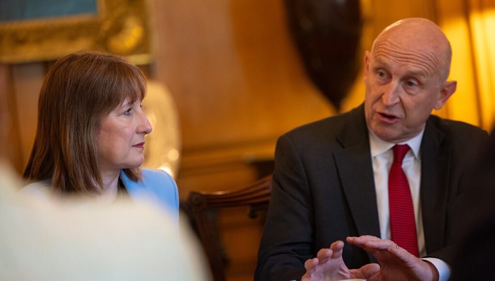 reeveshealey Two professionals sit at a meeting table, the woman in a light blue blazer listening as the man in a dark suit and red tie speaks gesturing with his hands.