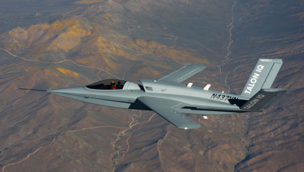 Gray stealth jet flying over arid desert mountains, cockpit visible and tail marked TALON Q with registration on fuselage