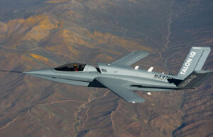Gray stealth jet flying over arid desert mountains, cockpit visible and tail marked TALON Q with registration on fuselage