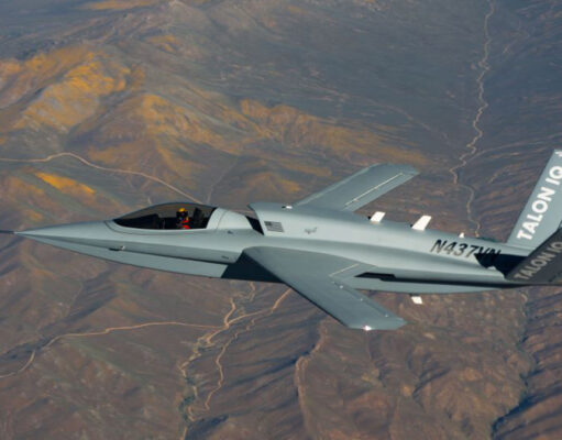 Gray stealth jet flying over arid desert mountains, cockpit visible and tail marked TALON Q with registration on fuselage