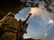 UK to award BAE Systems artillery components contract Soldier in camouflage with helmet operating a large mounted artillery gun, firing toward the sky from a trench-like position.