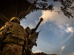 Soldier in camouflage with helmet operating a large mounted artillery gun, firing toward the sky from a trench-like position.