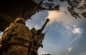 Soldier in camouflage with helmet operating a large mounted artillery gun, firing toward the sky from a trench-like position.
