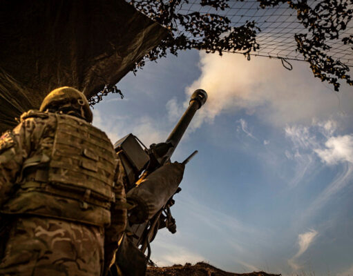 UK to award BAE Systems artillery components contract Soldier in camouflage with helmet operating a large mounted artillery gun, firing toward the sky from a trench-like position.