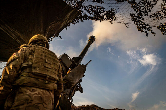 Soldier in camouflage with helmet operating a large mounted artillery gun, firing toward the sky from a trench-like position.