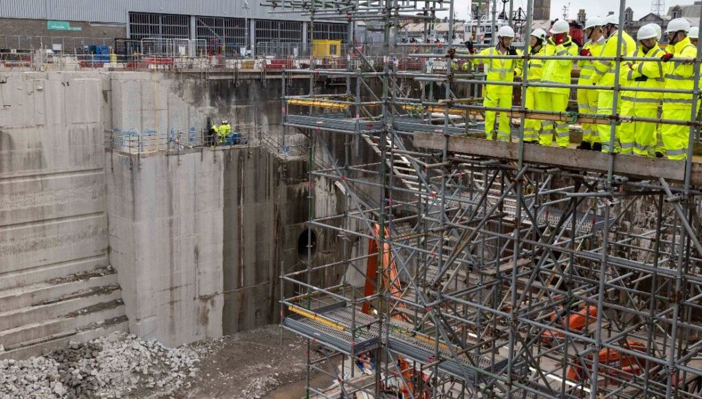 1779459659260 Construction site with workers in high-visibility suits and hard hats on scaffolding above a deep concrete excavation pit.