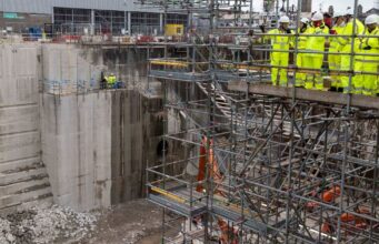First Sea Lord visits Babcock Devonport site to view work Construction site with workers in high-visibility suits and hard hats on scaffolding above a deep concrete excavation pit.
