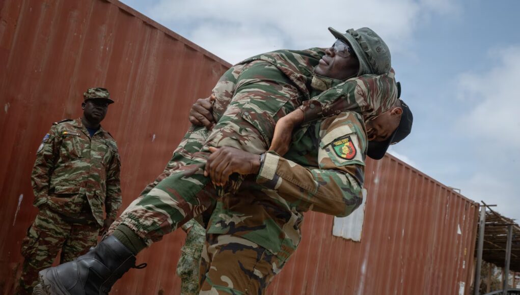 Two soldiers lift a bundled comrade in camouflage gear during a rescue or training exercise near a red shipping container outside.