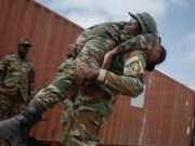 Two soldiers lift a bundled comrade in camouflage gear during a rescue or training exercise near a red shipping container outside.