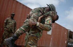 Two soldiers lift a bundled comrade in camouflage gear during a rescue or training exercise near a red shipping container outside.