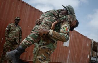 Two soldiers lift a bundled comrade in camouflage gear during a rescue or training exercise near a red shipping container outside.