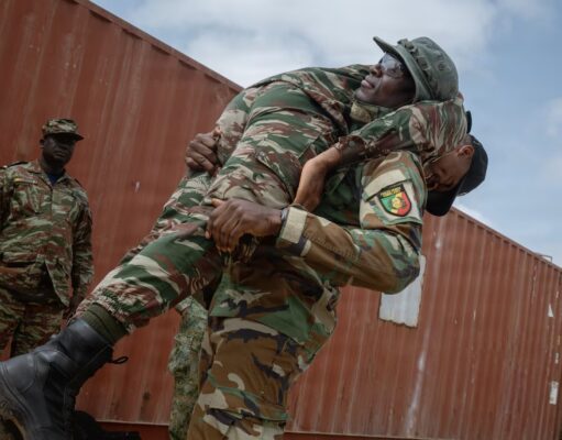 Two soldiers lift a bundled comrade in camouflage gear during a rescue or training exercise near a red shipping container outside.